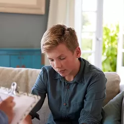 A happy family sitting together on a white sofa in a bright kitchen living area. The image shows two parents with their son and daughter, all laughing and enjoying each other's company. The mother has blonde hair, the father wears a red striped shirt, the son is in the middle with a gray shirt, and the teenage daughter sits across from them wearing a white top and blue jeans. The kitchen with white cabinets is visible in the background, creating a warm, homey atmosphere.